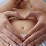 Zwei Hände formen liebevoll ein Herz um den Bauchnabel eines liegenden Babys – symbolisch für sanfte Nabelpflege und Babypflege.
