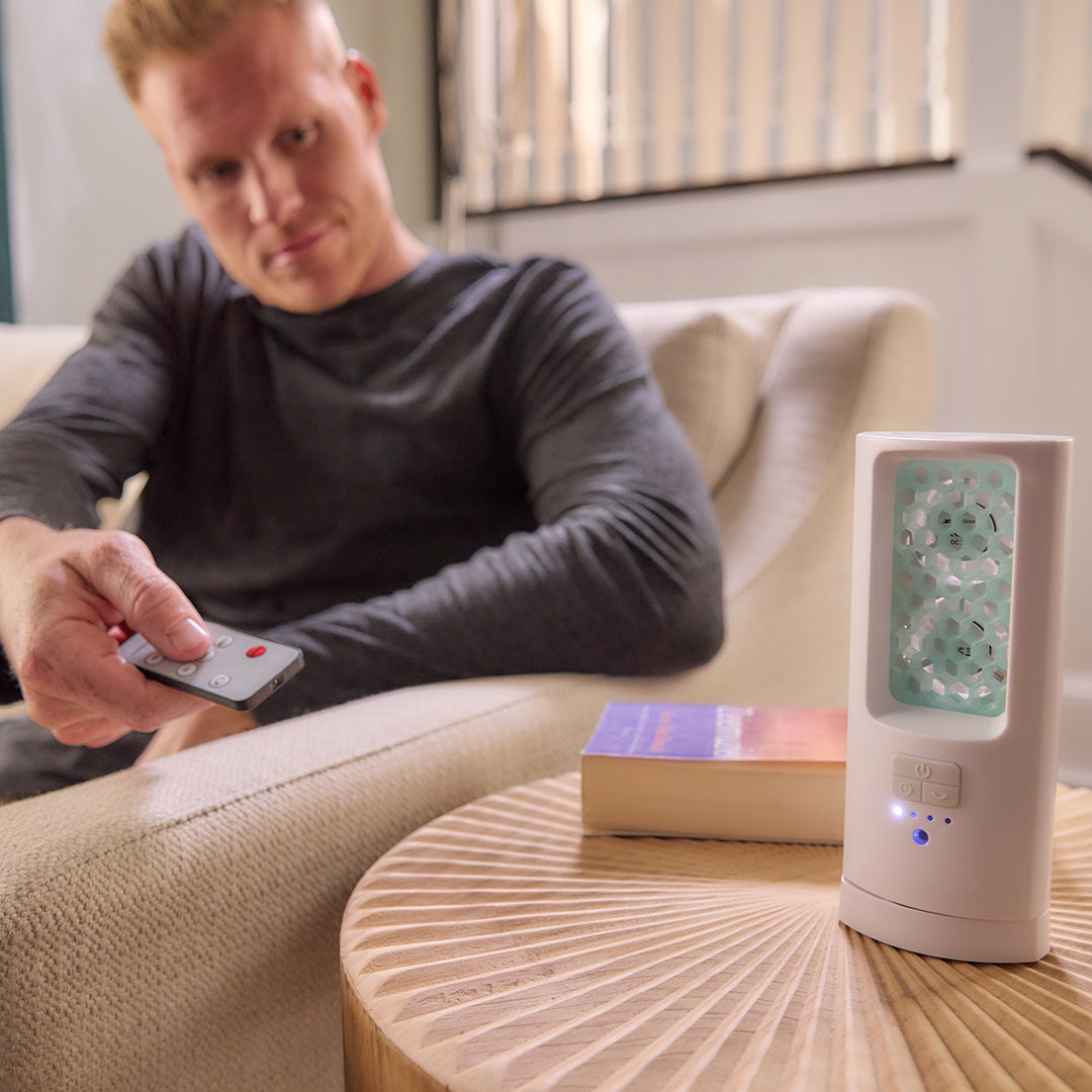 ScentDream Raumduftsystem mit eingesetztem Frischetraum-Pod auf einem Beistelltisch im Wohnzimmer, bedient von einem Mann auf dem Sofa per Fernbedienung.