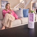 ScentDream Raumduftsystem auf einem Tisch mit eingesetztem violettem Lavendelglanz-Nachfüllpod; im Hintergrund sitzt eine entspannte Frau auf dem Sofa.