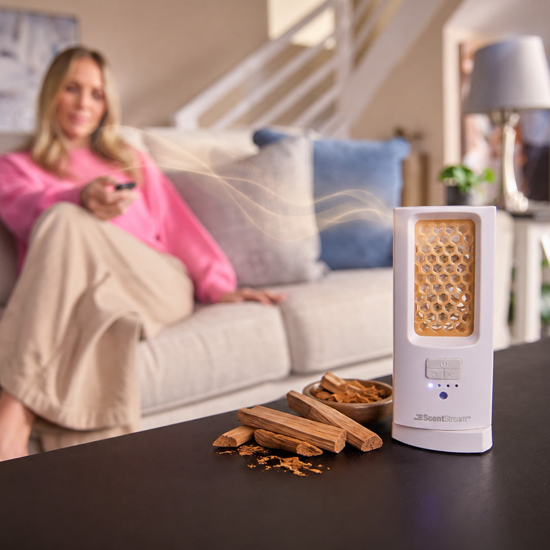 ScentDream Raumduftsystem auf einem Tisch mit eingelegtem Sandelholz-Duft-Pod, davor Sandelholzstücke und im Hintergrund eine entspannte Frau auf dem Sofa.