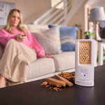 ScentDream Raumduftsystem auf einem Tisch mit eingelegtem Sandelholz-Duft-Pod, davor Sandelholzstücke und im Hintergrund eine entspannte Frau auf dem Sofa.
